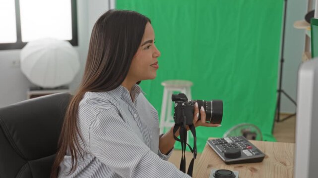 Woman holding camera and smiling while seated at an editing desk in a photo studio with green screen and softbox; creative concentration.