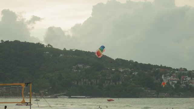 Parasail over island bay with colorful canopy and choppy sea, speedboat guiding passenger, dramatic cloudbank backdrop, tropical hillside resort visible, energetic tourist activity and winddriven