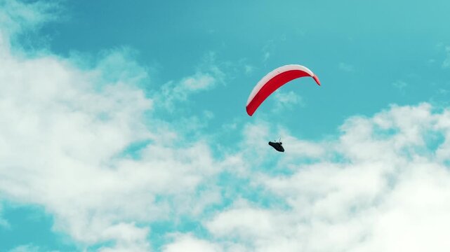 Paraglider with red canopy flying in blue sky with clouds. Solo paragliding flight with pilot silhouette suspended under wing, aerial sport activity.
