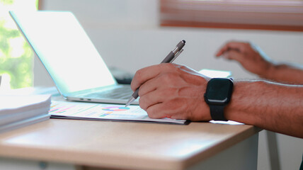 Fototapeta premium Close-up of professional analyzing business data, writing notes on report with pen while using laptop. Concept of data analysis, financial planning, productivity, and modern work.