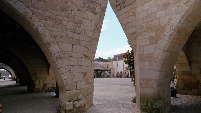 View of the central square and ancient architecture through a stone archway in Monpazier, Dordogne. Slow motion traveling shot.