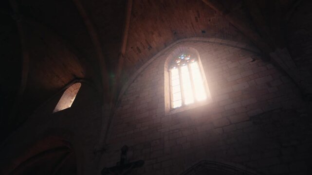 Sunbeams filter through a stone window inside a historic abbey in Monpazier, Dordogne, illuminating the vaulted ceiling. Slow motion traveling shot.