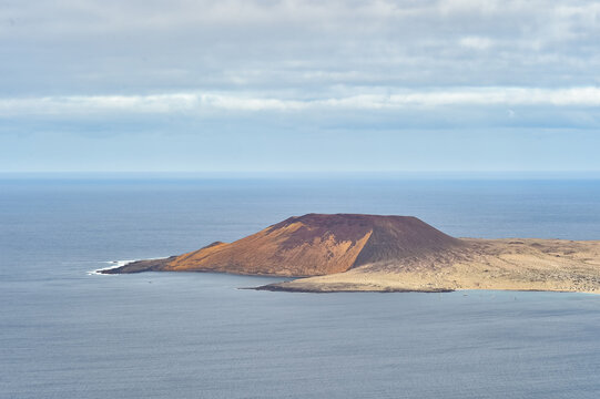 Volcanic island of La Graciosa seen from Lanzarote. Red and yellow volcanic crater cone surrounded by the Atlantic Ocean under a cloudy sky.