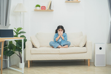 Fototapeta premium Young girl sitting on couch in living room, looking anxious and covering face with hands, expressing emotions of fear or worry in cozy home setting