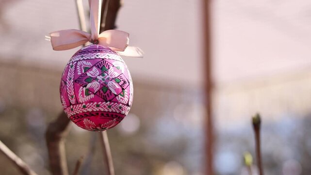 Close-up of a bright, traditionally painted Ukrainian pysanka (Easter egg) hanging on a tree branch against a blurred background. Decorative Easter egg suspended from a ribbon. Easter holiday