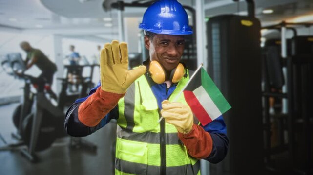 Construction worker man wearing blue hardhat and high visibility vest holds kuwait flag and raises open palm in gym; pride solidarity support.