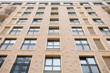Fototapeta premium Modern apartment building with warm brick facade. Uniform windows feature metal grilles and balconies. Rows of identical units create a rhythmic, orderly pattern. Ideal for urban architecture, housing