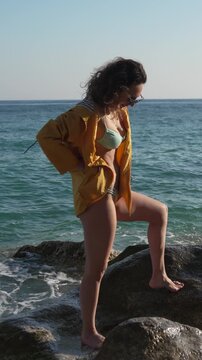 Woman beach ocean posing on rocky shore in yellow jacket while enjoying sunny day at the seaside