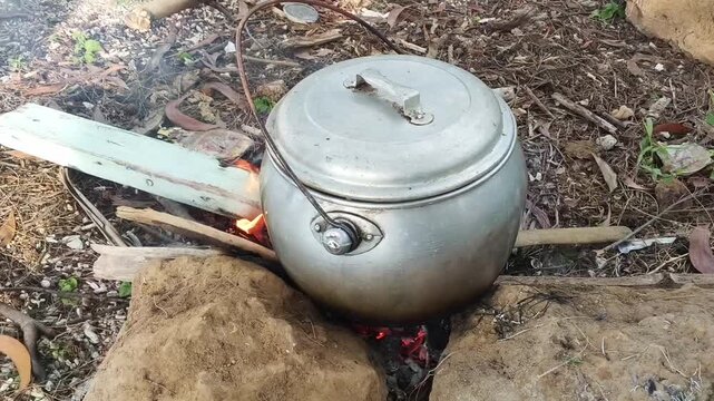 Traditional Cooking with Aluminum Pot over Open Wood Fire. A hand inserts a piece of firewood into a small open fire built between large rocks. A classic silver aluminum pot with a handle sits over th
