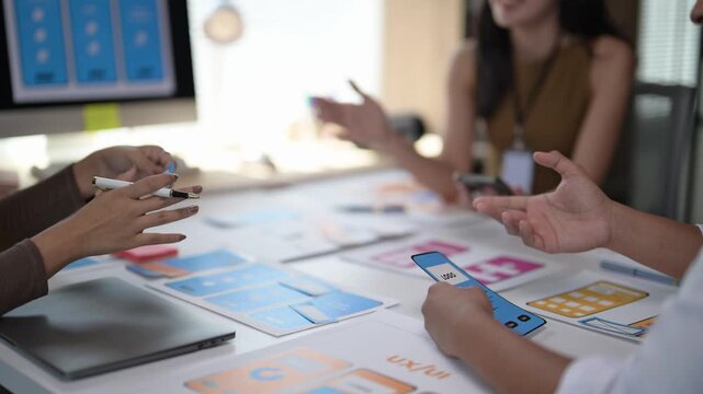 A group of people are sitting around a table with a white board and a laptop. They are working on a project and using pens and markers to write on the white board. Scene is collaborative and focused