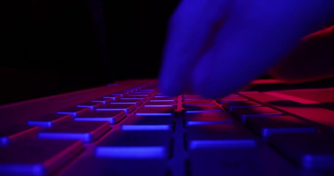 close up of fingers typing on computer keyboard in dark room by neon lights. cyber security concept