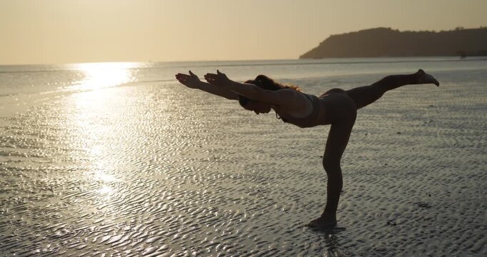 Silhouette of athletic woman shows single leg stand asana on sunrise ocean beach. Slim lady practices balance with physical activity against sun path on seaside
