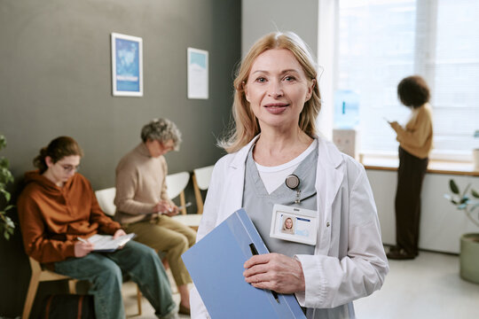 Portrait of middle aged Caucasian female doctor standing in clinic waiting room, holding patient files and smiling. Suitable for healthcare services, patient care, medical staffing promotion