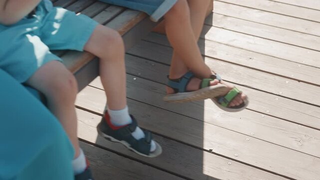 Kids feet and sandals on deck sunlit, two children sitting on bench with mixed sneakers and sandals, white socks and playful fidgeting, warm shadows across wooden planks, relaxed family weekend outing