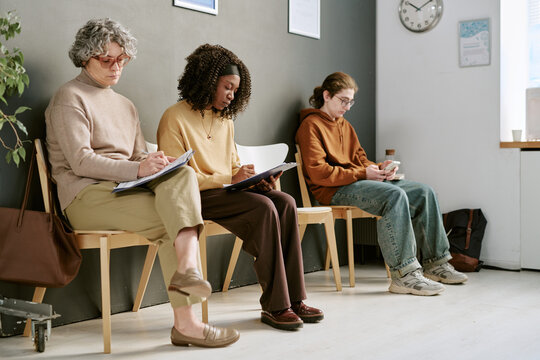Multiethnic women and teenager waiting, filling forms and checking smartphone in clinic lobby. Concept illustrating healthcare access, patient intake, insurance, medical services