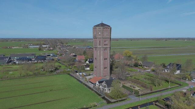 Aerial drone footage circling around a historic water tower while showing surrounding farmland, houses and countryside landscape from a wider perspective.