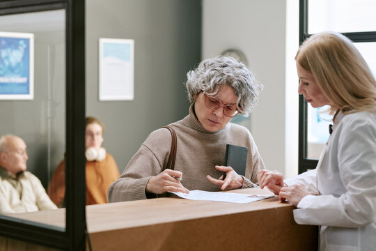 Senior Caucasian woman signing medical form while female doctor guiding paperwork at clinic reception, representing patient intake and healthcare administration. Useful for insurance, registration
