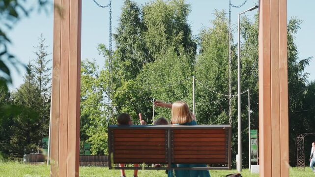 Parent and child on bench enjoying green park sunlight, wooden swing frame behind, gentle pointing gesture toward trees, relaxed weekend mood, leafy background and soft summer air creating warm family