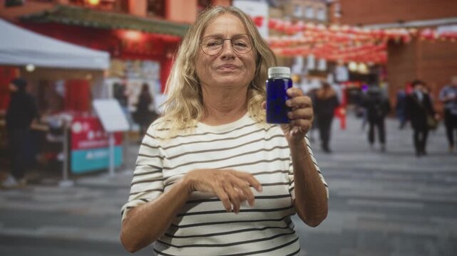 Senior blonde woman holding blue medicine bottle in hand while rubbing eye and squinting on crowded city street market; health concern.