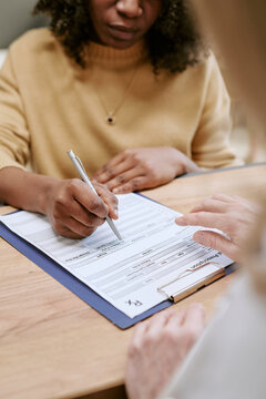 Vertical shot of Black woman signing medical form during consultation while clinician pointing at document. Supporting healthcare intake, patient registration, insurance or consent process