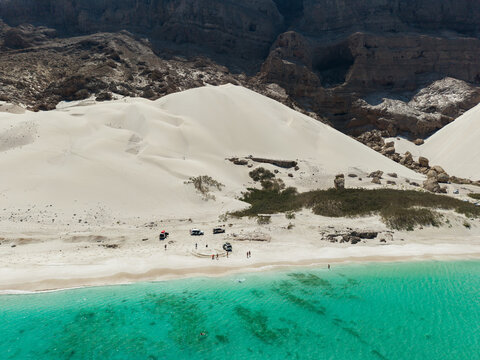 Aerial view of pristine white sand dunes meet the turquoise waters near the rugged mountains, creating a striking contrast of textures and tones, Erissel, Socotra, Yemen.