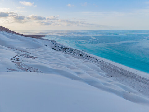 Aerial view of pure white sand dunes meet the turquoise sea under a pastel sky, creating a surreal coastal landscape, Erissel, Socotra, Yemen.