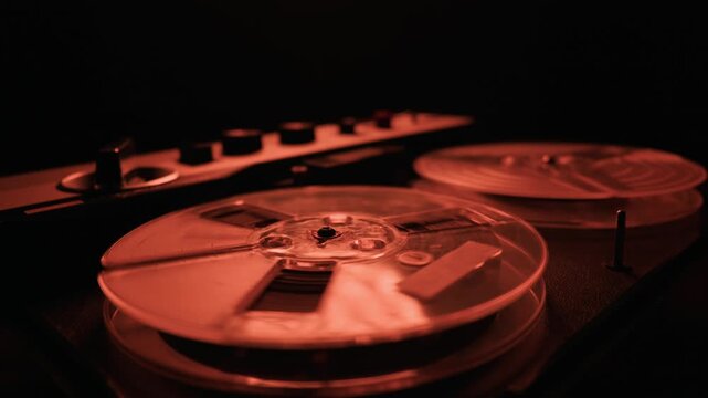 Vintage reel to reel audio tape recorder operates within a dark studio space. The transparent plastic reels spin continuously while illuminated by a red light. Analog sound equipment.