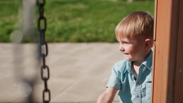 Family park weekend boy peeking playfully, caucasian child hides behind wooden post, giggle and quick peek, sunlight on hair, swing chain blurred foreground, grass and patio backdrop, energetic