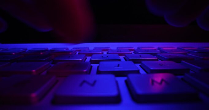 close up of fingers typing on computer keyboard in dark room by neon lights. cyber security concept
