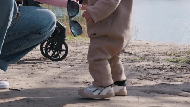 Unrecognizable toddler in a beige jumpsuit walks along a dirt path near a calm lake. Highlights the authentic concept of early childhood development, exploring nature, and learning.