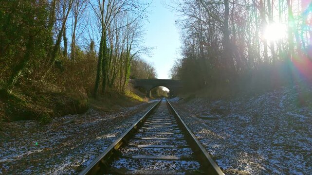 Freight train moving forward along railway tracks through forest cutting toward stone bridge first person view. Cargo convoy advancing straight ahead on rails across frosty ground between trees and