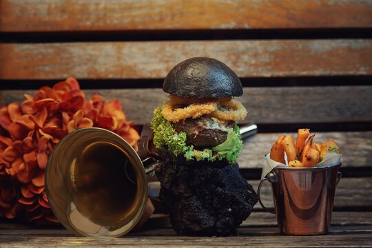 A striking black burger layered with crispy elements sits atop a dark stone base, accompanied by a side of golden fries in a small bucket. The rustic wood backdrop enhances the presentation