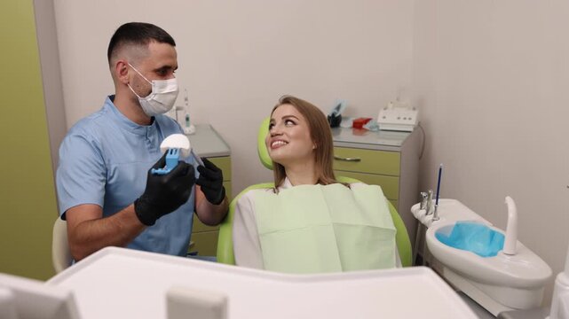 male doctor, patient attention, dental care. Dentist, man in face mask, is presenting dental model and tools to woman seated in dental chair, discussing her treatment options
