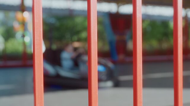Fenced bumper car ride park weekend distant view through red vertical bars, blurred bumper cars speeding around track, colorful canopies and greenery, candid spectator perspective, parent and child