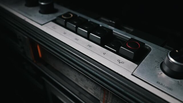 A human finger presses the black play button on a vintage silver cassette tape player. The retro audio system features classic mechanical switches and dials set against a dark background.