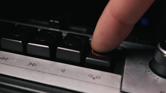 A person finger pushes the record button on a vintage silver cassette deck. Close up shot reveals the mechanical controls and classic audio equipment details.