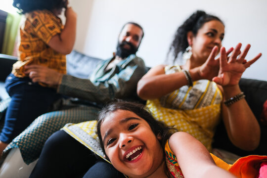 Happy female indian kid taking selfie with mobile phone camera at home with brother, mother and father in background - Family lifestyle concept - UGC wide lens content - Soft focus on child face