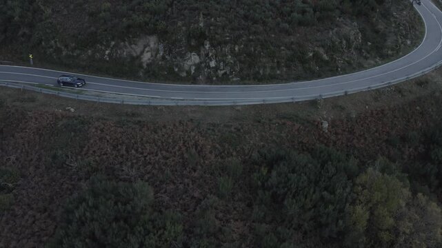 Black station wagon driving along mountain road in Serra da Estrela, Central Portugal, with shrub vegetation, pine patches and rocky hillside terrain in high-altitude landscape, drone tracking
