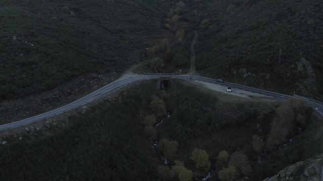N338 mountain road junction with stone culvert bridge over stream in Serra da Estrela, Central Portugal, showing wide valley, vehicles, riparian trees and vegetation in rugged terrain, drone pull away