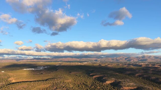 Drone view captures a vast pinyon covered mesa landscape at sunrise. Patches of light highlight the rugged terrain as the drone flies toward distant mountains under a beautiful cloudy blue sky.