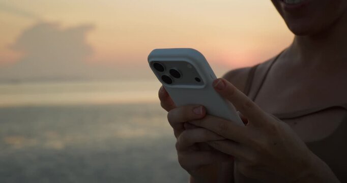 Crop view of woman hands write message on smartphone as stands on sunset beach. Young female uses mobile phone and rests after yoga session on evening coast