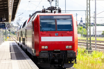 Fototapeta premium Red regional train locomotive stand platform german railway station overhead catenary wires tracks. Passenger rail transport infrastructure commuter mobility travel connection europe sunny summer day