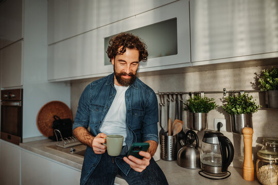 Man enjoying morning coffee scrolling on smartphone in kitchen