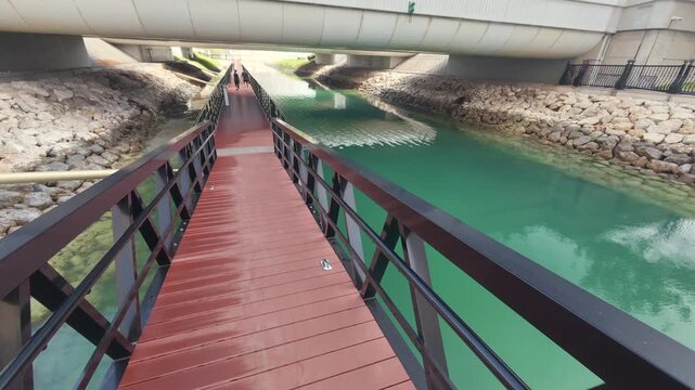Red wooden walkway leads under a concrete bridge. Green river flows with stone banks on both sides. Three keyframes capture different perspectives of the scene. No people are present
