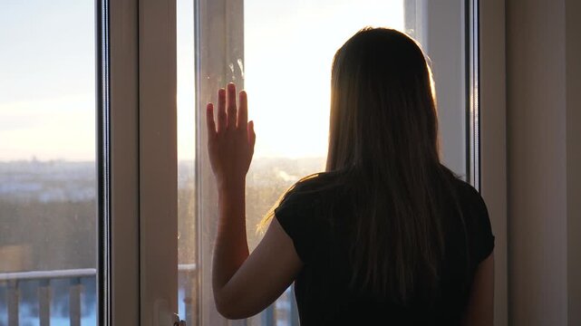 Solitary woman standing by window. Female looking at city during beautiful sunset. People feeling loneliness and sadness while staying at home, representing quarantine and self isolation