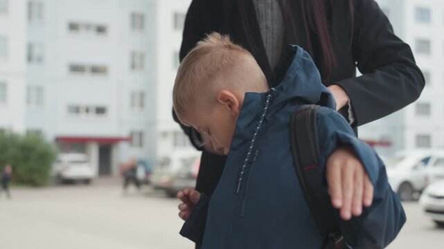 Young white boy with backpack being helped by mother at apartment entrance, jacket hood adjusted, reluctant expression, urban morning, preparing for school, subtle anxiety and parental care captured