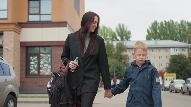 Smiling parent and child at school, front view with adult holding hand and carrying bag and bottle, boy in jacket walking confidently past modern building and parked cars, warm glance and casual