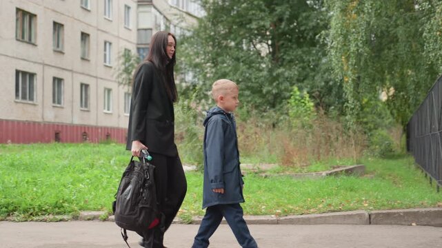 White mother escorting son school gate, apartment block backdrop with greenery, mother carrying backpack while son opens iron gate, moment of reassurance and last minute comfort before class