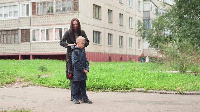 White mother adjusts son backpack school, tender zipper check and gentle hug, apartment and greenery backdrop, confident child stepping toward classroom, warm farewell and parental support