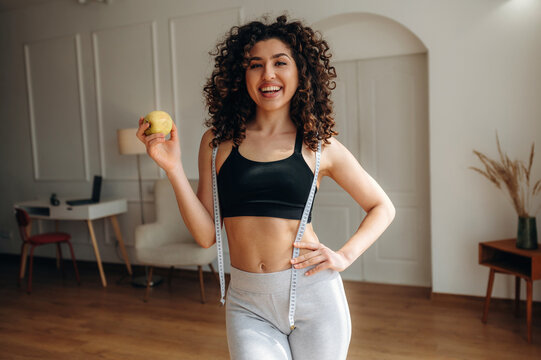Young woman with curly hair holds an apple while wearing a black sports bra and gray leggings, standing in a bright indoor space with wooden flooring and minimal decor